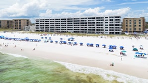 Un hotel frente a la playa con varios pisos, una playa de arena con sombrillas azules y un cielo despejado.