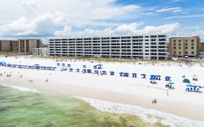 A beachfront hotel with multiple floors, a sandy beach in front, and several blue umbrellas. - Surf Dweller 202 - Emerald Shores (Fort Walton Beach)