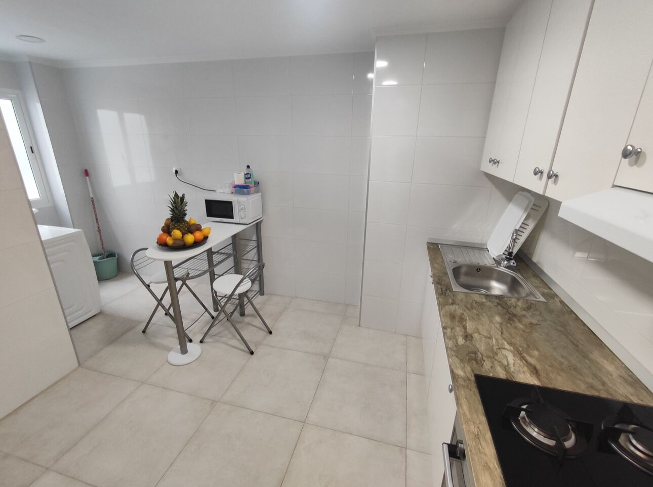 A compact kitchen with white cabinets, a small dining table with chairs, a fruit bowl, and a sink.