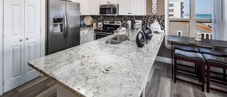 Una cocina moderna con armarios blancos, encimera de granito, electrodomésticos de acero inoxidable y vista a la playa.