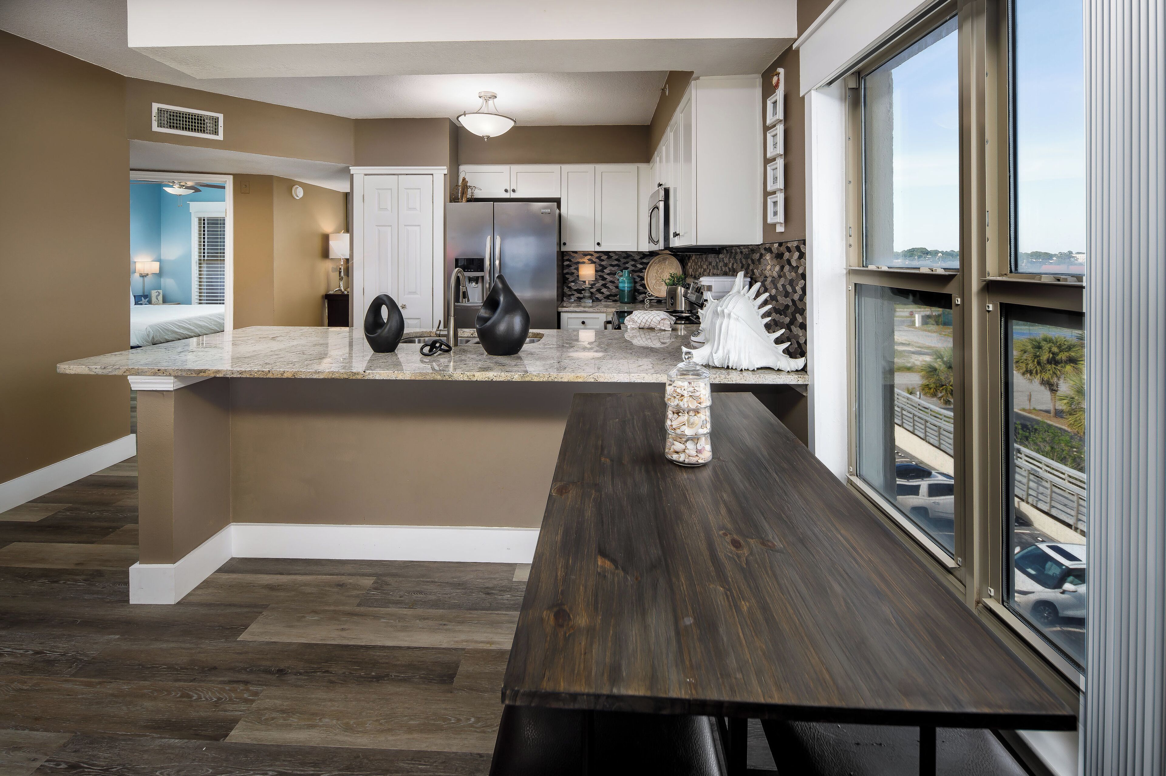 A modern kitchen with a marble countertop, a stainless steel refrigerator, and a dining area with a bench and a view through large windows.