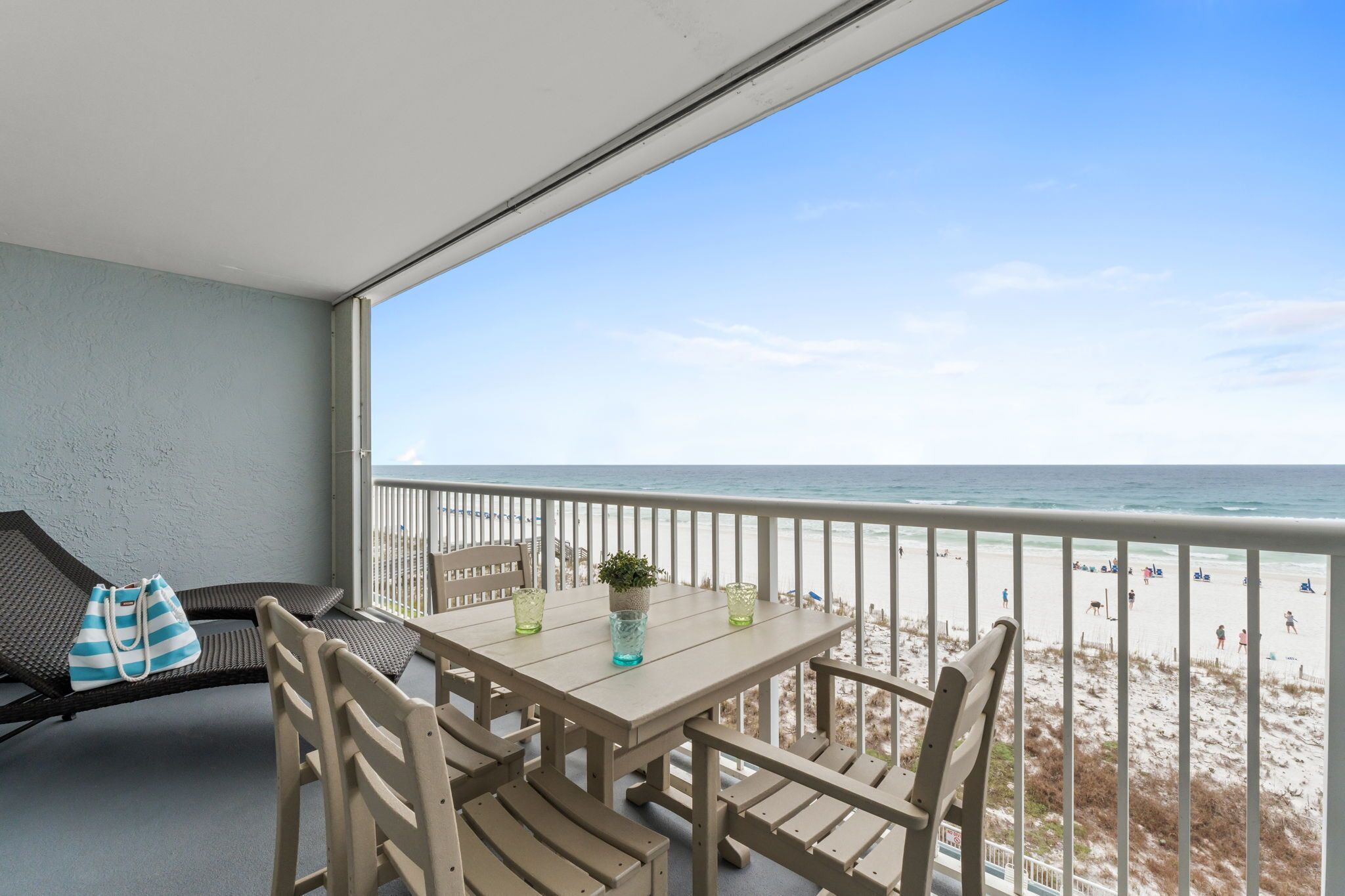 A balcony with a table and chairs, overlooking a beach with people and a clear sky.