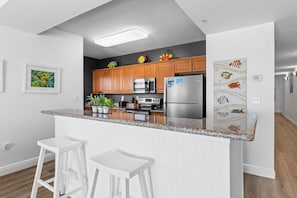 A modern kitchen with wooden cabinets, stainless steel appliances, a granite countertop, and white bar stools. - Pelican Isle 613 - Salty Sunsets (Fort Walton Beach)