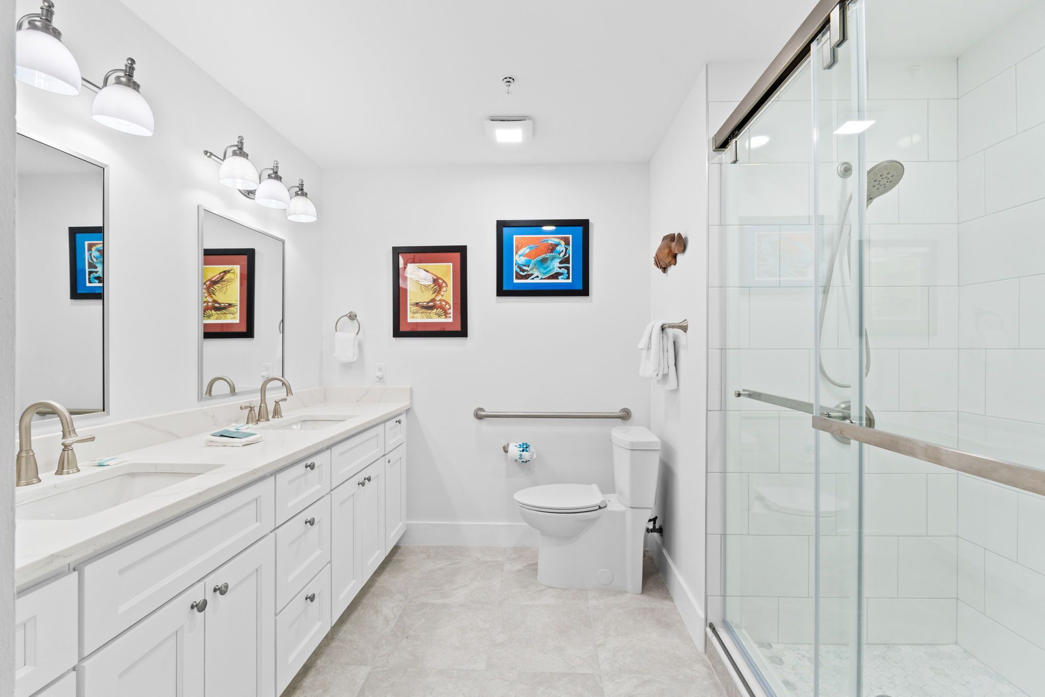 A modern bathroom with a white sink, vanity, and toilet. There are framed pictures on the wall, a glass shower enclosure, and a handrail.