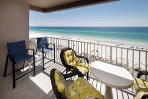 A balcony with wicker chairs and a table, overlooking a beach with umbrellas and people. - Island Princess 701 - Beach Bum Bliss (Fort Walton Beach)