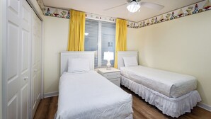 A bedroom with two beds, a nightstand, a lamp, a window with yellow curtains, and a ceiling fan. - Island Princess 701 - Beach Bum Bliss (Fort Walton Beach)