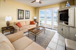 A living room with a sofa, coffee table, and TV, a balcony with a view of the ocean, and a sailboat decoration. - Sea Dunes 504 - Vacation Vibe (Fort Walton Beach)