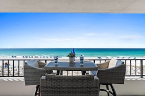 Un balcón con sillas de mimbre y una mesa, con vista a una playa con sombrillas y un cielo azul despejado.