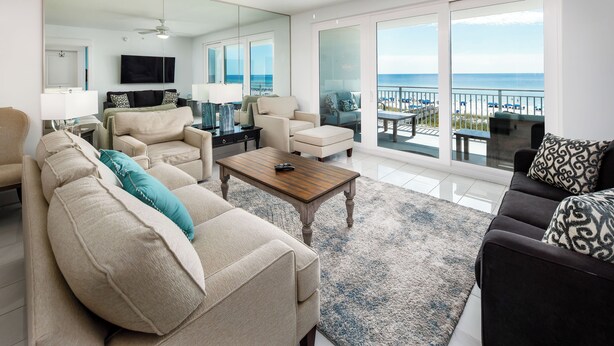 A spacious living room with a large mirror, a ceiling fan, and a view of the ocean through large sliding glass doors. - Blu 306 - The Gulf Jewel (Fort Walton Beach)