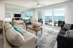A spacious living room with a large mirror, a ceiling fan, and a view of the ocean through large sliding glass doors. - Blu 306 - The Gulf Jewel (Fort Walton Beach)