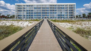 A multi-story hotel with a central courtyard, a wooden boardwalk leading to the entrance, and a sandy beach area. - Blu 306 - The Gulf Jewel (Fort Walton Beach)