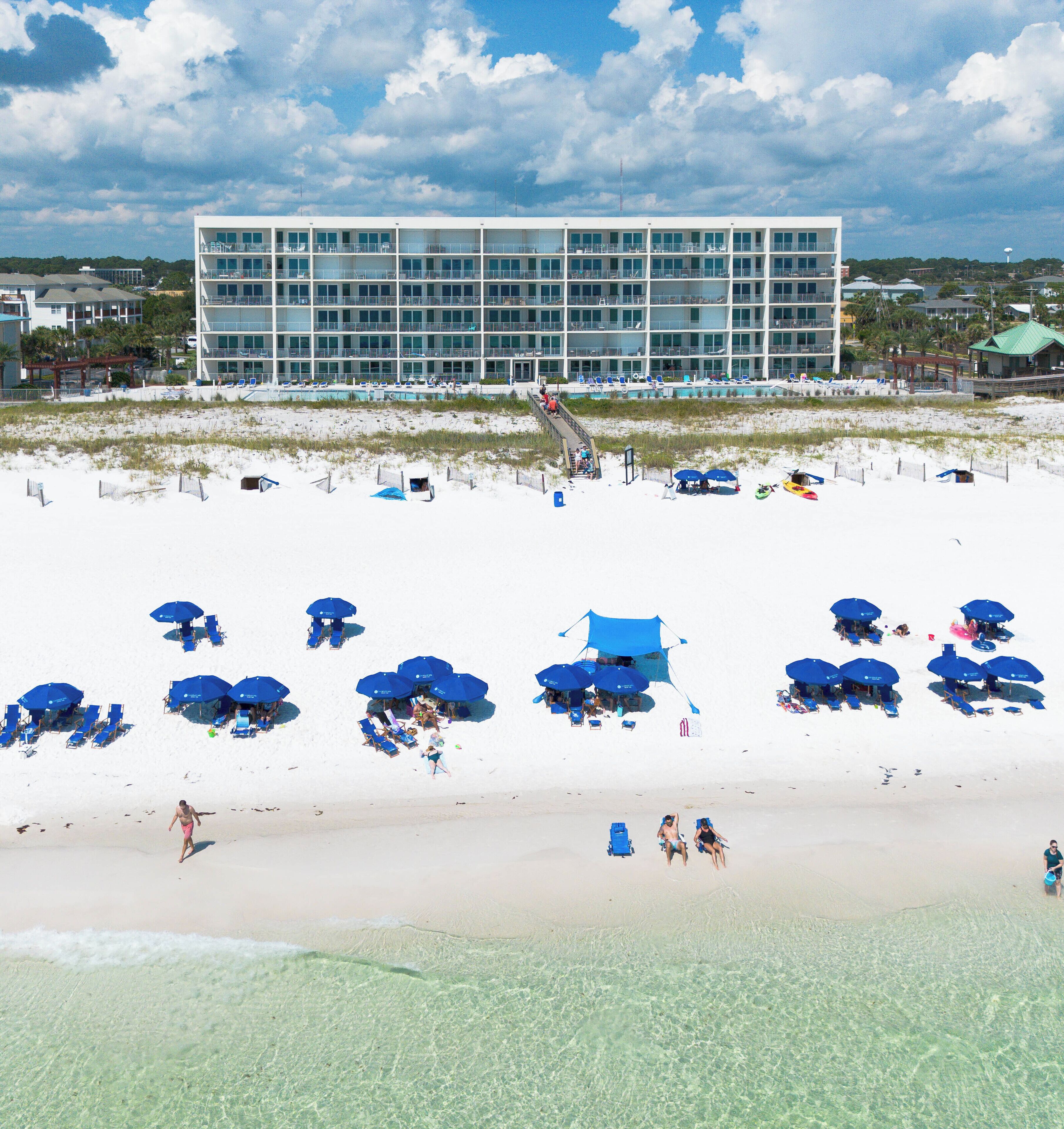 A beach with clear shallow waters, a large building with multiple balconies, and several blue umbrellas.