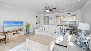 A modern living room with a large flat-screen TV, a ceiling fan, and a kitchen in the background. - Gulfside 401 - Sunny Side Up (Fort Walton Beach)