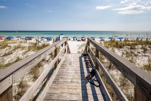 Ein hölzerner Steg führt zu einem Strand mit Sonnenschirmen und Menschen.
