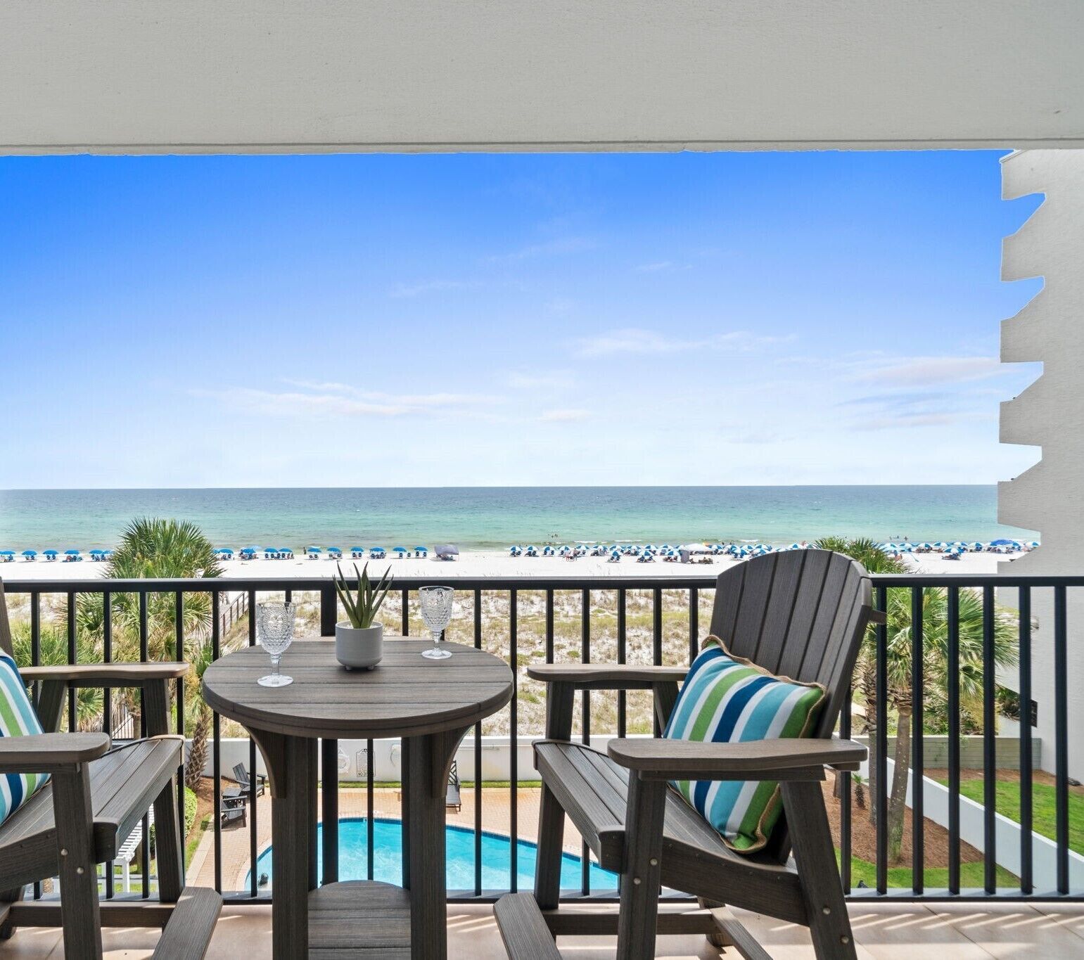 A balcony with a view of the ocean, a pool, and beach umbrellas.
