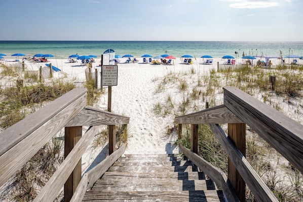 A wooden staircase leading to a sandy beach with blue umbrellas and beach chairs. - Dune Pointe 402 - Shells, Surf & Sea Turtles (Fort Walton Beach)