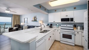 A modern kitchen with white cabinets, a large island with a sink, and a view of the ocean through a balcony. - Pelican Isle 307 - Sunkissed & Vibing (Fort Walton Beach)