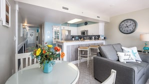 A modern kitchen with white cabinets, a stainless steel refrigerator, and a microwave. There is a dining area with a glass table and chairs, and a gray sofa with a decorative pillow.