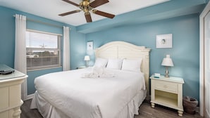 A bedroom with a large bed, a ceiling fan, a nightstand with a lamp, and a window with blinds.