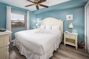 Une chambre à coucher avec un grand lit, une table de chevet, une commode, un ventilateur de plafond et une fenêtre avec des stores.