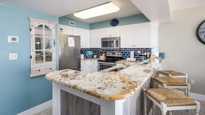 A kitchen with white cabinets, a stainless steel refrigerator, a microwave, and a granite countertop. There are three bar stools with woven seats.