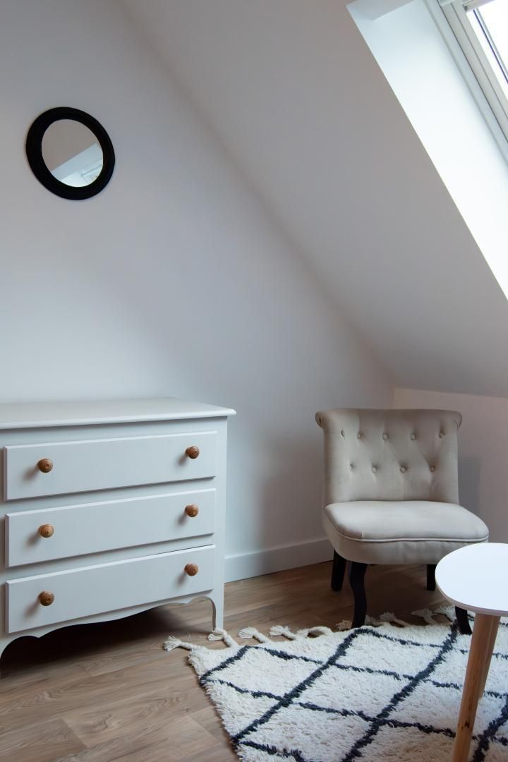 A room with a white dresser with wooden knobs, a tufted armchair, a round mirror on the wall, and a small round table.