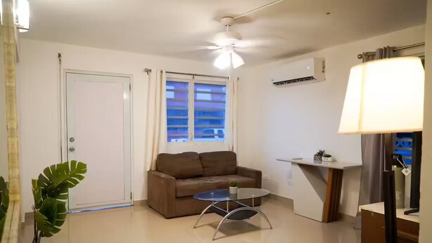 A modern living room with a brown sofa, a glass coffee table, a ceiling fan, a window with blinds, and a small plant. - Cozy 3BR + Parking, Near Airport — Solar Backup! (Carolina)