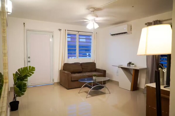 A modern living room with a brown sofa, a glass coffee table, a ceiling fan, a window with blinds, and a small plant.
