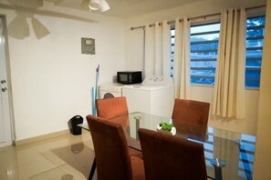 A small, modern room with a glass dining table, two brown chairs, a microwave, a washing machine, and a small plant on the table.