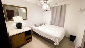 A small, clean room with a bed, a bedside table with a lamp, a mirror, a ceiling fan, and a window with curtains. - Cozy 3BR + Parking, Near Airport — Solar Backup! (Carolina)