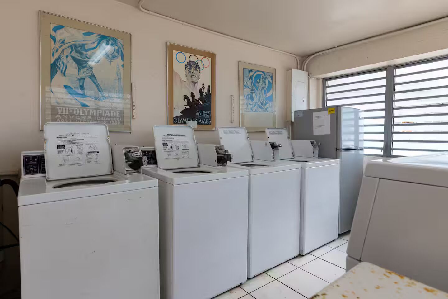 A laundry room with multiple washing machines and dryers, posters on the wall, and a window with blinds.