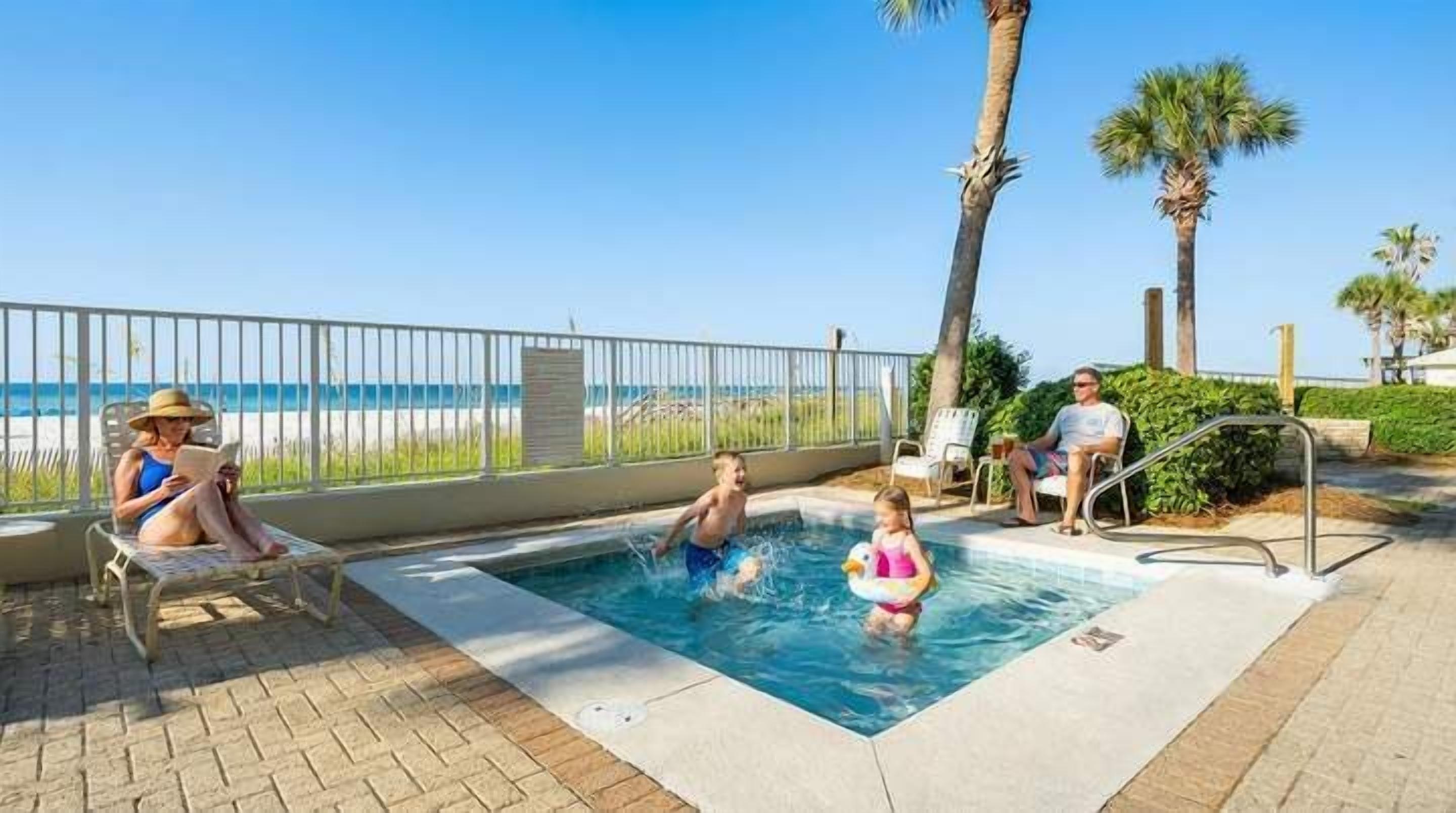A swimming pool area with a view of the beach, a fenced deck, and palm trees.