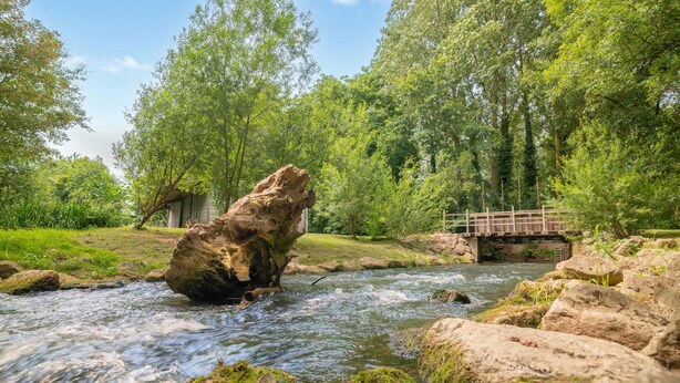 A river flowing through a forest with a wooden bridge and a rustic building in the background. - Ancien moulin restauré (Clerques)