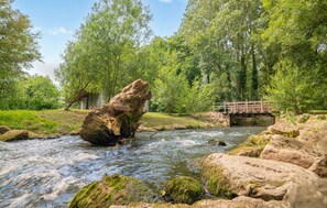 Une rivière traverse une forêt, avec un pont en bois et un bâtiment rustique en arrière-plan.
