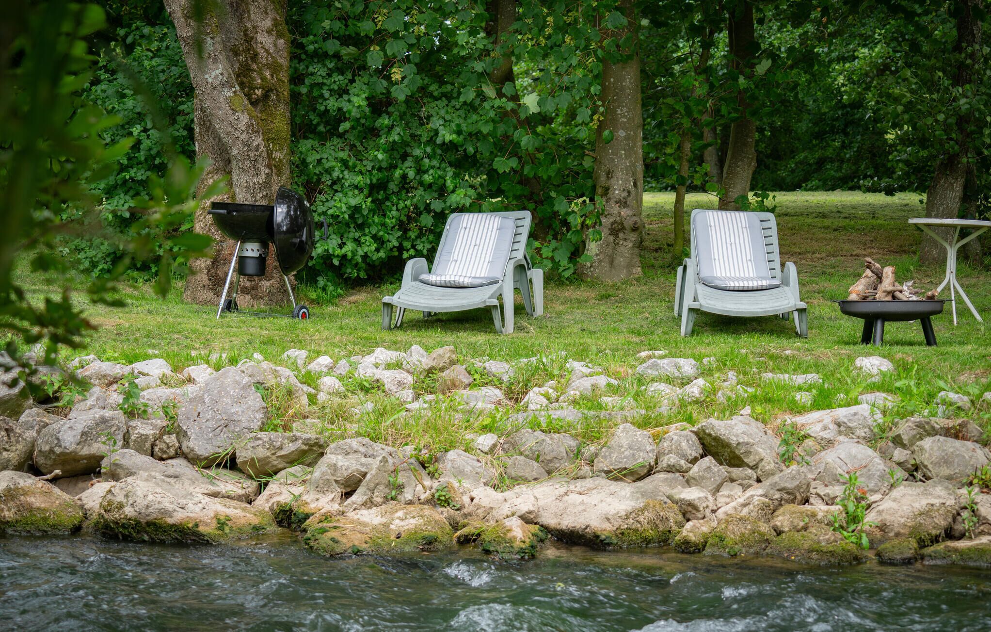A riverside setting with two lounge chairs and a grill, surrounded by rocks and greenery.