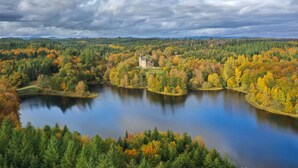 Ein großes Gewässer, umgeben von dichtem Wald mit Herbstlaub, ein markantes Gebäude in der Mitte und ein bewölkter Himmel.