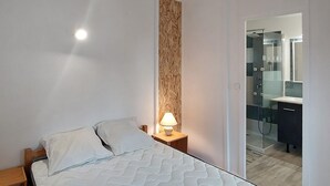 Ein Schlafzimmer mit einem Bett, einem Nachttisch mit Lampe und Blick ins Bad mit Dusche und Spiegel.