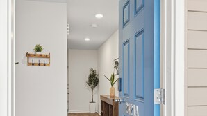 Ein Flur mit einer blauen Tür, weißen Wänden, Holzboden, einer Pflanze und einem kleinen Regal.