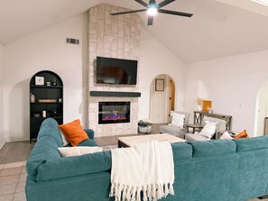 Un salon doté d’une cheminée en pierre, d’un meuble de rangement noir pour appareils électroniques, de canapés turquoise et d’un ventilateur de plafond.