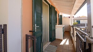 Un pasillo estrecho con contraventanas verdes, un escalón que lleva a un balcón y suelo de baldosas.