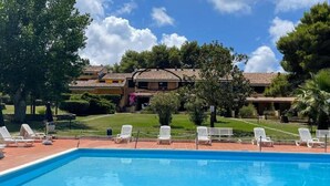 Una piscina con lettini, circondata da un prato curato e un edificio con tetto in tegole.