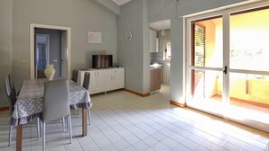 Uma área de refeições com mesa e cadeiras, uma cozinha ao fundo e uma porta de vidro deslizante que dá para uma varanda.