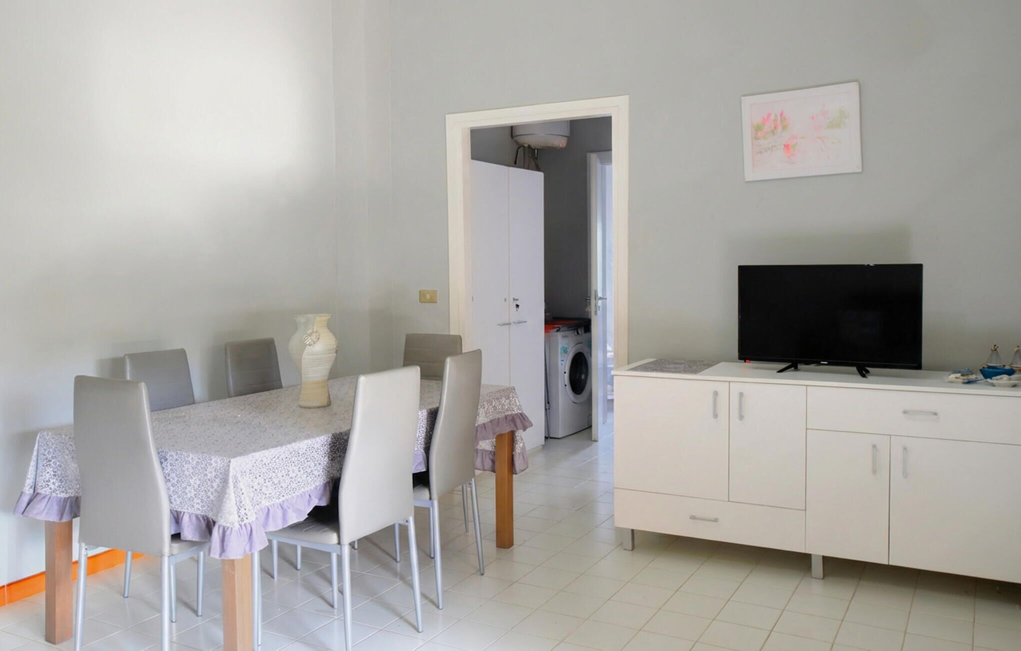 Un comedor con una mesa dispuesta para cuatro personas, un televisor en un mueble y una lavadora al fondo.