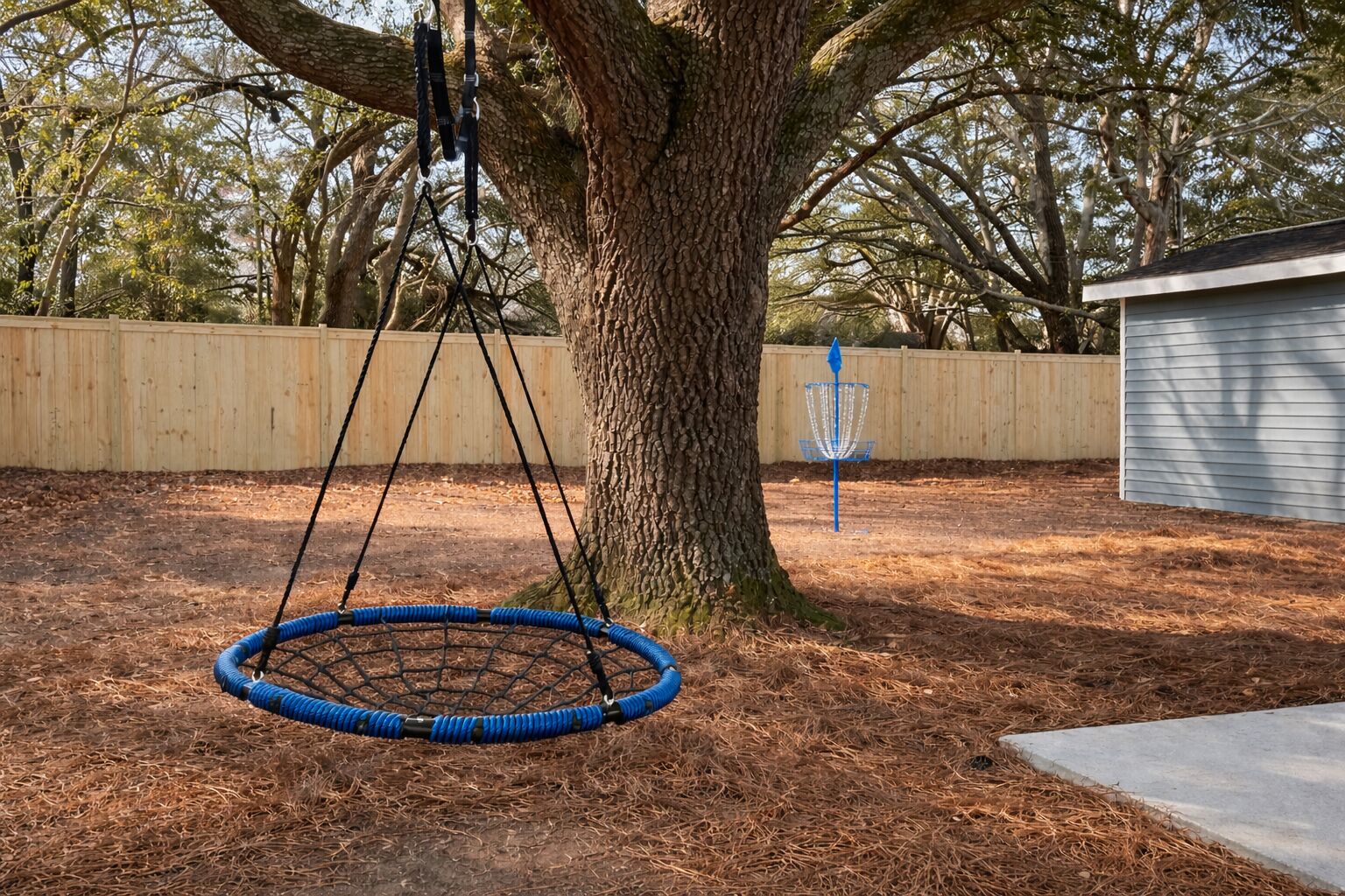 Een achtertuin met een schommel in een boom, een frisbee-basket en een houten omheining.
