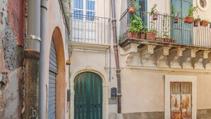 Eine schmale Straße mit einem Gebäude, das eine grüne Tür, einen Balkon mit Blumentöpfen und ein verrostetes Metallgitter aufweist.