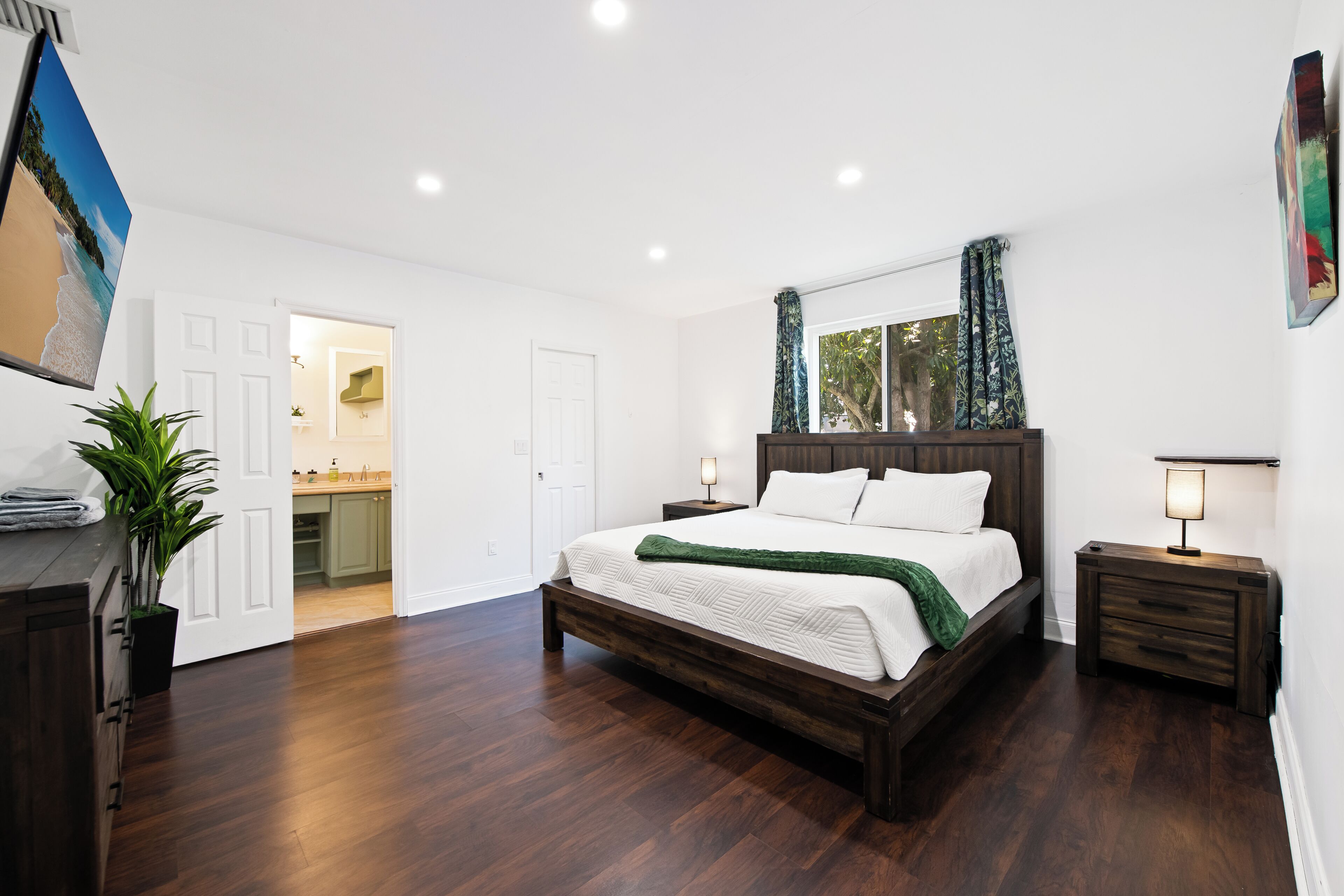 A bedroom with a wooden bed, white walls, a large window with curtains, and a view of a kitchen and bathroom.