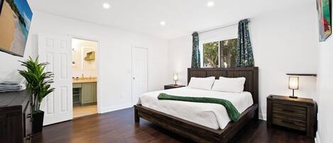 A bedroom with a wooden bed, white walls, a large window with curtains, and a view of a kitchen and bathroom.
