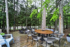 An outdoor patio with a round wooden table surrounded by wicker chairs, set against a backdrop of tall trees. - Torch + Timber House (Rapid City)