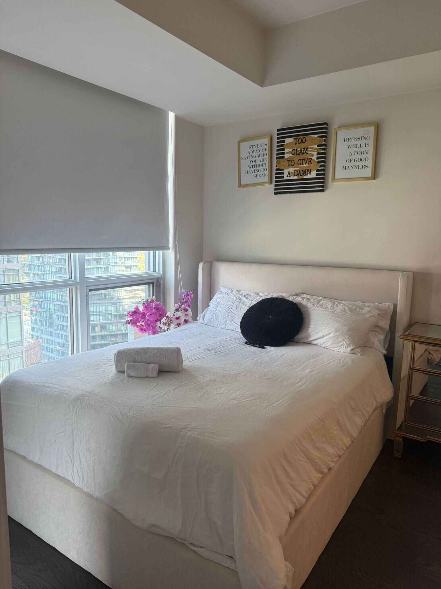 A neatly made bed with a white comforter and pillows, a small bedside table with a lamp, and a window with a view of buildings.