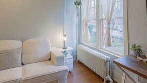 Een kamer met een witte bank, een klein nachtkastje met een lamp, een radiator en een raam met gordijnen.